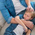 child with gastroenteritis or the stomach flu sleeping in dads lap on the couch